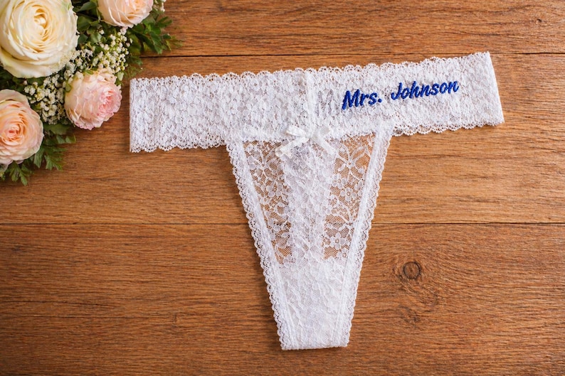 May include: White lace thong with the embroidered text "Mrs. Johnson" in blue. The lingerie has a delicate floral pattern and a small bow. The thong is displayed on a wooden surface, with a bouquet of roses in the background.