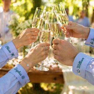 May include: Light blue long-sleeved shirts with embroidered names and small icons on the cuffs. The cuffs feature the names Olivia, Sophia, William, and James in green thread, accompanied by small flower, star, and leaf motifs, while holding champagne flutes.