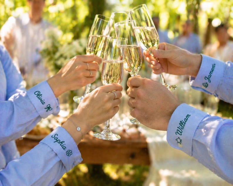 May include: Light blue long-sleeved shirts with embroidered names and small icons on the cuffs. The cuffs feature the names Olivia, Sophia, William, and James in green thread, accompanied by small flower, star, and leaf motifs, while holding champagne flutes.