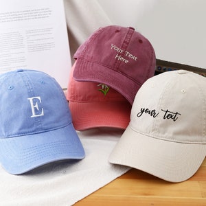 Könnte beinhalten: Vier verschiedene Baseballkappen mit gestickten Designs. Eine ist blau mit einem weißen Buchstaben E, eine ist beige mit schwarzem kursivem Text, der "your text" sagt, eine ist rot mit einer weißen Blume und eine ist burgunderrot mit weißem Text, der "Your Text Here" sagt.