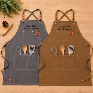 May include: Two canvas aprons, one gray and one brown, featuring cross-back straps with leather accents. Each apron has a chest pocket, a large front pocket holding a whisk, wooden spoon, and spatula, and the embroidered text Head Chef Kris J. Petersen.