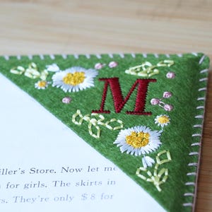 May include: Green felt bookmark with embroidered white daisies and leaves. The letter "M" is embroidered in red in the centre of the bookmark.