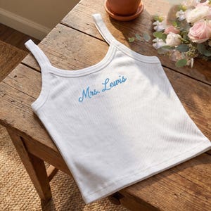 May include: White ribbed tank top with thin straps. The text "Mrs. Lewis" is embroidered in light blue across the chest. The top is lying on a wooden surface, with a small pot and flowers in the background.
