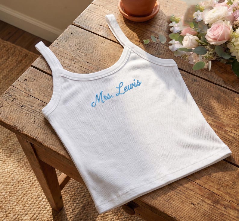 May include: White ribbed tank top with thin straps. The text "Mrs. Lewis" is embroidered in light blue across the chest. The top is lying on a wooden surface, with a small pot and flowers in the background.