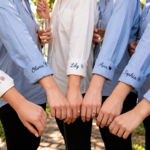 May include: A group of long-sleeved button-down shirts in light blue and white, featuring rolled-up cuffs embroidered with names and small icons. The names Isla, Olivia, Lily, Ava, Sophia, and Chloe are stitched in dark thread on the cuffs.