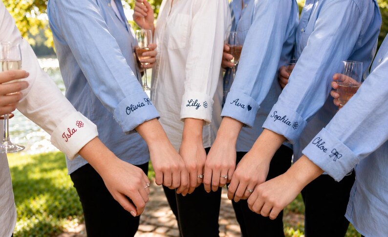 May include: A group of long-sleeved button-down shirts in light blue and white, featuring rolled-up cuffs embroidered with names and small icons. The names Isla, Olivia, Lily, Ava, Sophia, and Chloe are stitched in dark thread on the cuffs.