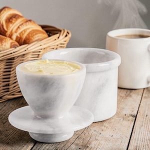 Könnte beinhalten: Ein weißer Butterbehälter aus Marmor mit Deckel, gefüllt mit gelber Butter, steht auf einer Holzoberfläche. Ein Korb mit Croissants und eine weiße Kaffeetasse sind im Hintergrund. Der Butterbehälter hat einen Durchmesser von ca. 10 cm.
