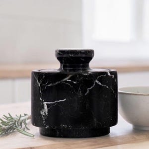 Può includere: Un contenitore in marmo nero con coperchio, con venature bianche. Il corpo cilindrico poggia su una piccola base. Un rametto di rosmarino e una ciotola bianca sono sullo sfondo. Il contenitore è probabilmente per uso in cucina.