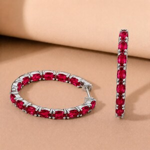 May include: A pair of hoop earrings crafted from silver-toned metal, each featuring a continuous row of vibrant, oval-cut red gemstones set in a prong setting. One earring is displayed lying flat in a circular shape, while the other stands upright.