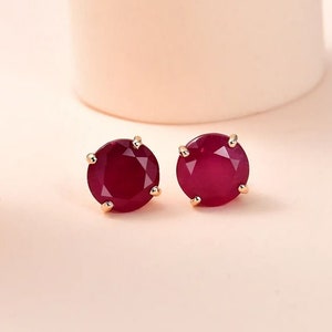 May include: A pair of gold stud earrings with a round, red gemstone in each earring.