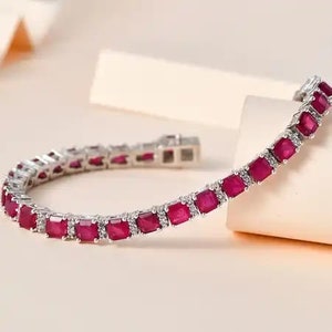 Könnte beinhalten: Ein silbernes Armband mit quadratisch geschliffenen, tiefroten Edelsteinen. Jeder Edelstein ist mit kleinen, klaren Akzentsteinen besetzt. Das Armband ist gebogen, der Verschluss ist am Ende sichtbar. Der Schmuck wird auf einer hellen Oberfläche präsentiert.