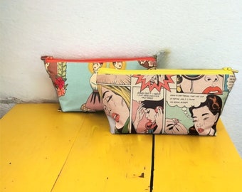 Può includere: Due pochette con cerniera colorate con illustrazioni in stile fumetto. La pochette superiore ha uno sfondo blu con una donna in un vestito rosso e un design floreale. La pochette inferiore ha uno sfondo verde con un'illustrazione in stile fumetto di donne con fumetti. Il testo nei fumetti dice "SICK OUT! - AND I JUST GOT MY HAIR DONE HALF WAY!" e "MAN, I'VE THROWN THAT LAST CUP OF COFFEE AND I. I. I. I'M GOING ANOTHER!"