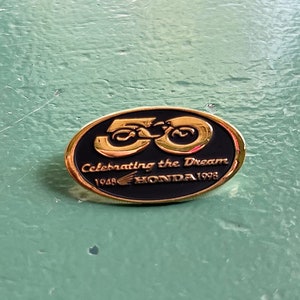 May include: A gold-colored oval pin with a black background. The pin features the number "50" in gold with a stylized design. The text "Celebrating the Dream 1948 HONDA 1998" is printed in gold on the black background.