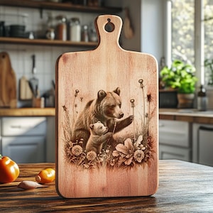 May include: Wooden cutting board with a handle, featuring a detailed illustration of a mother bear and cub in a field of flowers and tall grass. The board is made of light-colored wood and is suitable for kitchen use.