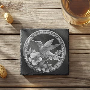May include: A square, black slate coaster featuring an engraved hummingbird design, encircled by floral elements and a decorative frame. The coaster rests on a wooden surface, with peanuts and a glass of liquid nearby.