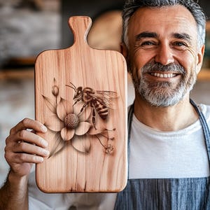 Può includere: Tagliere in legno con manico, con un'incisione dettagliata di un'ape su un fiore. La tavola ha una venatura naturale del legno e un colore marrone chiaro. Il design dell'ape e del fiore aggiunge un elemento decorativo.