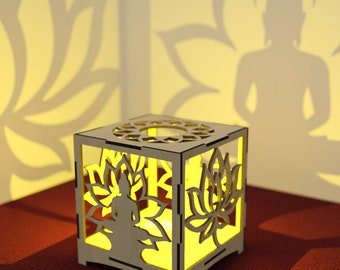 Peut inclure: Un bougeoir en bois avec un motif découpé représentant une figure méditant entourée de fleurs de lotus. Le bougeoir est éclairé de l'intérieur, projetant une lueur chaleureuse.