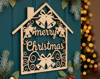 Puede incluir: Decoración navideña de madera con forma de casa con las palabras "merry Christmas" recortadas. La decoración presenta diseños de campanas y remolinos. El fondo es una puerta verde azulado con vegetación navideña y campanas doradas.