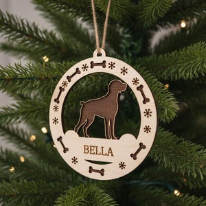 Peut inclure: Ornement de Noël en bois avec une silhouette marron d'un chien dans un cadre circulaire. Le cadre est orné d'os et de flocons de neige découpés. Le nom "BELLA" est gravé en bas. Une boucle en ficelle est attachée pour accrocher.