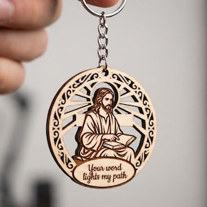 Puede incluir: Llavero de madera con un diseño cortado con láser de Jesús leyendo un libro, rodeado de un patrón decorativo de rayos de sol. El llavero incluye el texto "Your word lights my path" en un óvalo. Unido a un llavero plateado.
