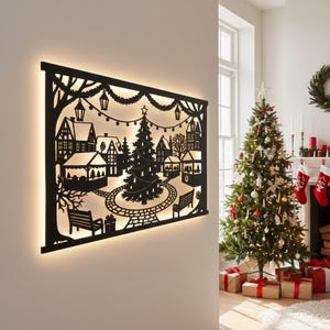 Könnte beinhalten: Schwarze Weihnachts-Dorf-Wandkunst aus Metall mit beleuchtetem Hintergrund. Die Silhouette zeigt eine festliche Szene mit einem Weihnachtsbaum, Gebäuden, Bänken und Lichterketten. Das Kunstwerk ist an einer weißen Wand befestigt.