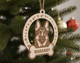 Può includere: Ornamento natalizio in legno con un disegno di pastore tedesco. L'ornamento circolare ha un'etichetta a forma di osso con la scritta "BARANBY". È decorato con impronte di zampe e fiocchi di neve e pende da uno spago.