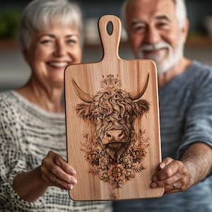 May include: A wooden chopping board with a hand-burned design of a Highland cow with floral accents. The board has a handle and is being held by two people.