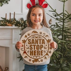 Op de afbeelding: Een rond houten bord met de tekst "SANTA STOP HERE" in een bruin lettertype. Het bord is versierd met kerstthema-illustraties, waaronder rendieren, snoepstokken en cadeaus. Een jong persoon houdt het bord voor een kerstboom.