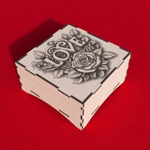 Puede incluir: Una caja de madera con tapa con bisagras. La tapa está decorada con una ilustración en blanco y negro de una rosa y la palabra "LOVE" en una fuente estilizada.