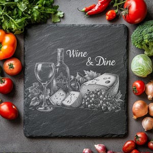 Puede incluir: Tabla de servir de pizarra negra con el texto "Wine & Dine" grabado en blanco. La tabla presenta un dibujo de una botella de vino, una copa de vino, queso, uvas y hojas.