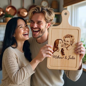 May include: A light-coloured wooden chopping board with a handle. The board has a laser-etched portrait of a couple and the names "Mithew & Julia" in script. The couple is laughing. The board is held by a person.