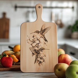 Può includere: Un tagliere in legno con manico presenta un design inciso a laser di due colibrì in volo con accenti floreali.