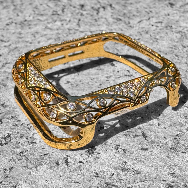 May include: Gold-tone watch case with a decorative, intricate design featuring clear rhinestones. The case is designed to fit a square-shaped watch face.