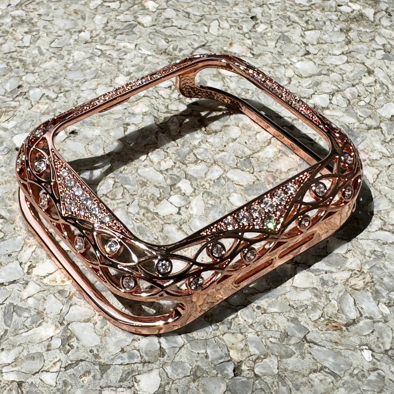 May include: Rose gold Apple Watch case with a clear back and a decorative, intricate design featuring clear rhinestones.