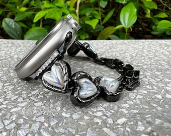 May include: A black and silver watch band with a heart-shaped design. The band is made of metal and has a silver finish. The hearts are iridescent and have a pearlescent finish.