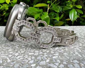 May include: A silver-toned smartwatch with a decorative band. The band features a series of interlocking geometric shapes, each outlined with sparkling clear crystals. The watch and band are resting on a textured gray surface, with green foliage in the background.