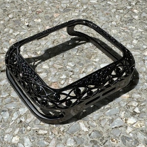 May include: Black metal Apple Watch case with a decorative, intricate design. The case has a black diamond-like finish.