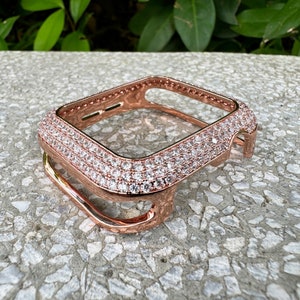 Pode incluir: Uma capa protetora para smartwatch em ouro rosa com uma estrutura retangular incrustada com inúmeras pequenas pedras claras e brilhantes na superfície superior, com recortes precisos para os botões laterais do dispositivo.