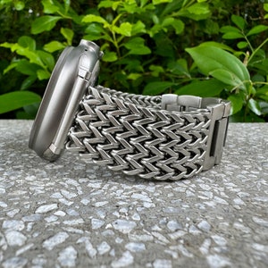 Könnte beinhalten: Ein silbernes Metallarmband mit Fischgrätenmuster. Das Armband besteht aus kleinen, ineinandergreifenden Gliedern, die ein flexibles und robustes Design schaffen.