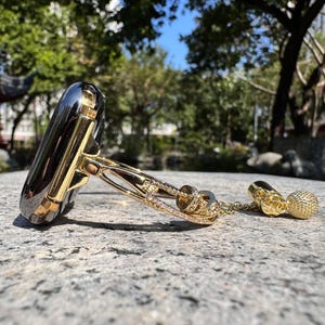Op de afbeelding: Een gouden en zwarte horlogeband met een decoratieve ketting en een kleine gouden hanger. De band is gemaakt van metaal en heeft een diamant-achtig patroon.
