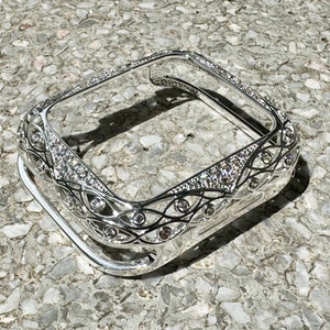May include: A silver-toned Apple Watch case with a decorative, intricate design featuring a filigree pattern and clear rhinestones.