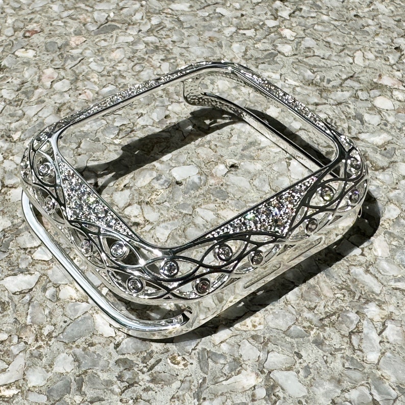 May include: A silver-toned Apple Watch case with a decorative, intricate design featuring a filigree pattern and clear rhinestones.
