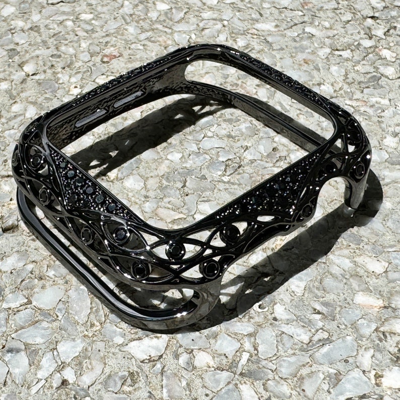 May include: Black metal Apple Watch case with a decorative, intricate design featuring black rhinestones.