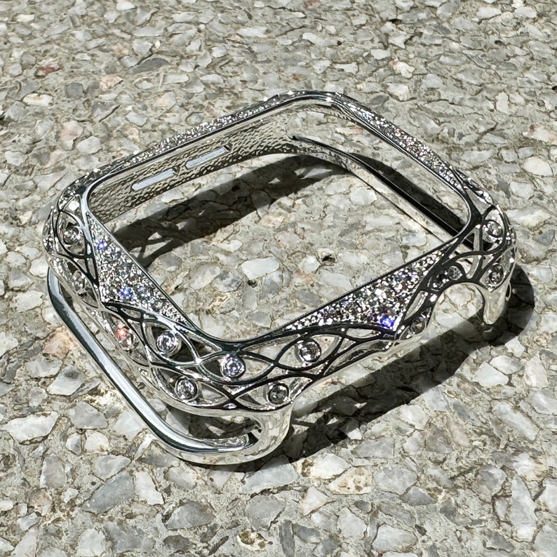 May include: A silver-toned Apple Watch case with a decorative, intricate design featuring a filigree pattern and clear rhinestones.
