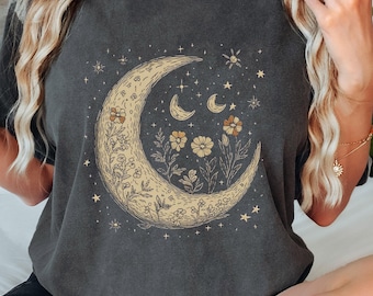 May include: A dark gray t-shirt with a crescent moon and floral design. The moon is outlined in black and filled with a light yellow color. The flowers are in shades of yellow and orange.