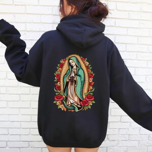 May include: Black hoodie featuring a colorful graphic of the Virgin Mary surrounded by roses. The artwork is centered on the back of the hoodie. The Virgin Mary is depicted with a teal cloak and a white dress, with her hands clasped in prayer.