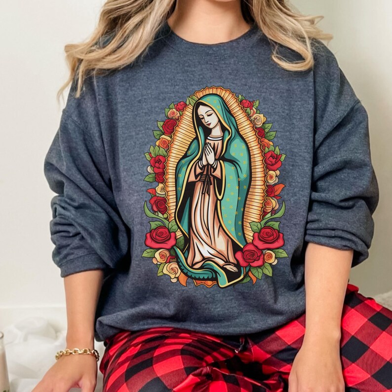 May include: A dark gray sweatshirt featuring a graphic of the Virgin Mary surrounded by roses. The illustration depicts Mary in a teal robe and white dress, with hands clasped in prayer. The design is framed by a golden halo and a wreath of red and yellow roses.