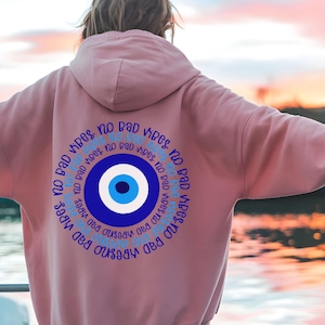 May include: A light pink hoodie with a circular design on the back. The design features a blue and white eye symbol surrounded by the words "NO BAD VIBES" in a blue, circular pattern. The hoodie has a hood and long sleeves.