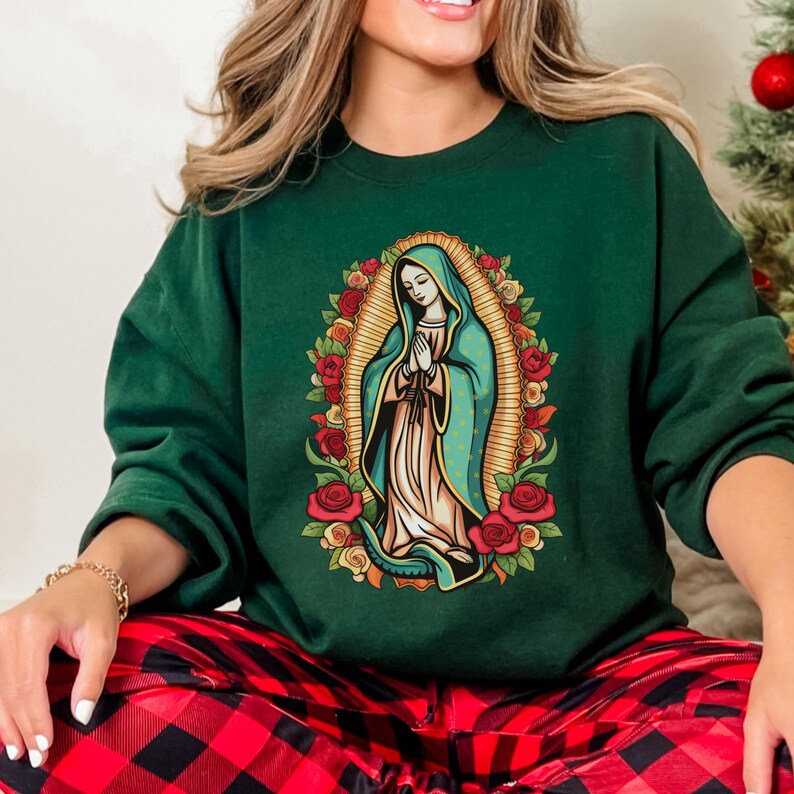 May include: A dark green sweatshirt featuring an illustration of the Virgin Mary surrounded by red roses. The Virgin Mary is depicted in a blue robe with hands clasped in prayer. The sweatshirt is a crew neck style.