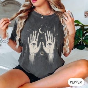 Może przedstawiać: Szary t-shirt z nadrukiem graficznym przedstawiającym dwie dłonie z oczami, p&oacute;łksiężyc i gwiazdy. T-shirt ma luźny kr&oacute;j i okrągły dekolt.